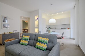 Ein modernes Wohnzimmer mit einem grauen Sofa, zwei gemusterten Kissen, einem Essbereich mit weißem Tisch und Stühlen sowie einer Küche mit weißen Schränken und einer Pendelleuchte.