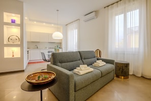 Ein modernes Wohnzimmer mit einem grauen Sofa, einem kleinen runden Tisch mit einer Schüssel und einer Küche im Hintergrund.
