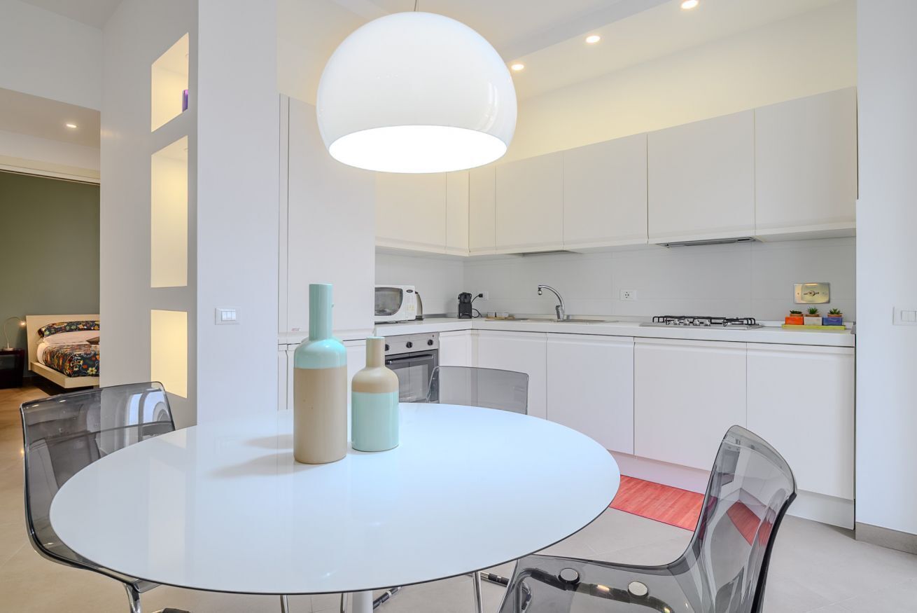 Une cuisine moderne avec des meubles blancs, une table ronde en verre et un luminaire suspendu.