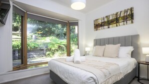 A bedroom with a large bed, a view of greenery through a sliding door, and a painting on the wall.
