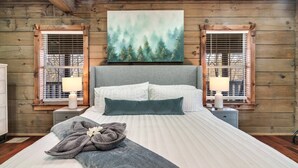 A bedroom with a large bed, two bedside tables with lamps, a painting of a forest, and wooden walls. - 🏡 Luxury Smoky Mountain Retreat | Mega Spa, Game Room & Stunning Views (Cosby)