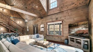 A spacious living room with a high ceiling, wooden walls, a large window with a view of a river, a staircase, and a television on a wooden stand. - 🏡 Luxury Smoky Mountain Retreat | Mega Spa, Game Room & Stunning Views (Cosby)