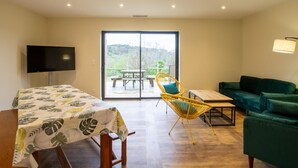 A room with a bed, a TV, a table, a chair, a sofa, and a sliding glass door leading to an outdoor area.