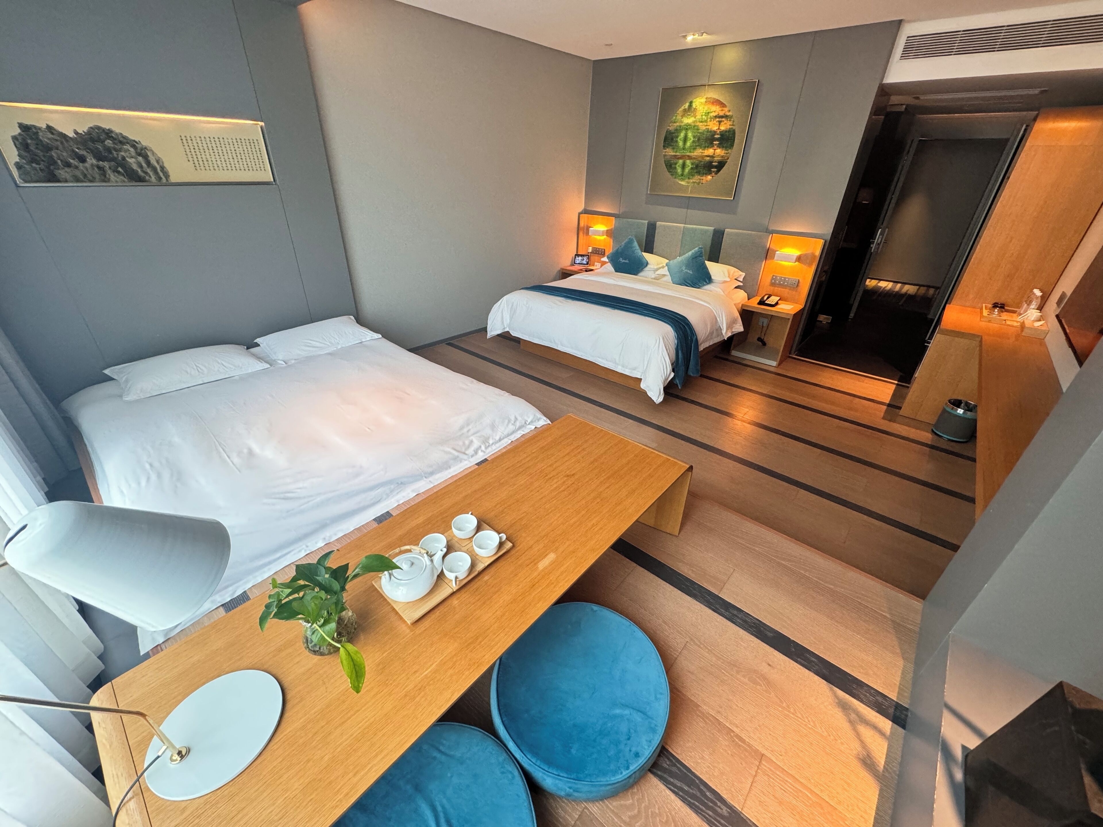 A modern hotel room with two beds, a wooden desk with a teapot and cups, a blue cushioned stool, and a small plant.