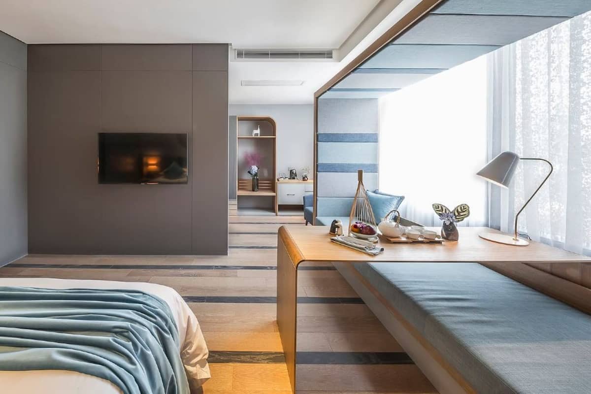 Ein modernes Hotelzimmer mit einem großen Bett, einem Schreibtisch mit Lampe, einem an der Wand befestigten Fernseher und einem Fenster mit transparenten Vorhängen.