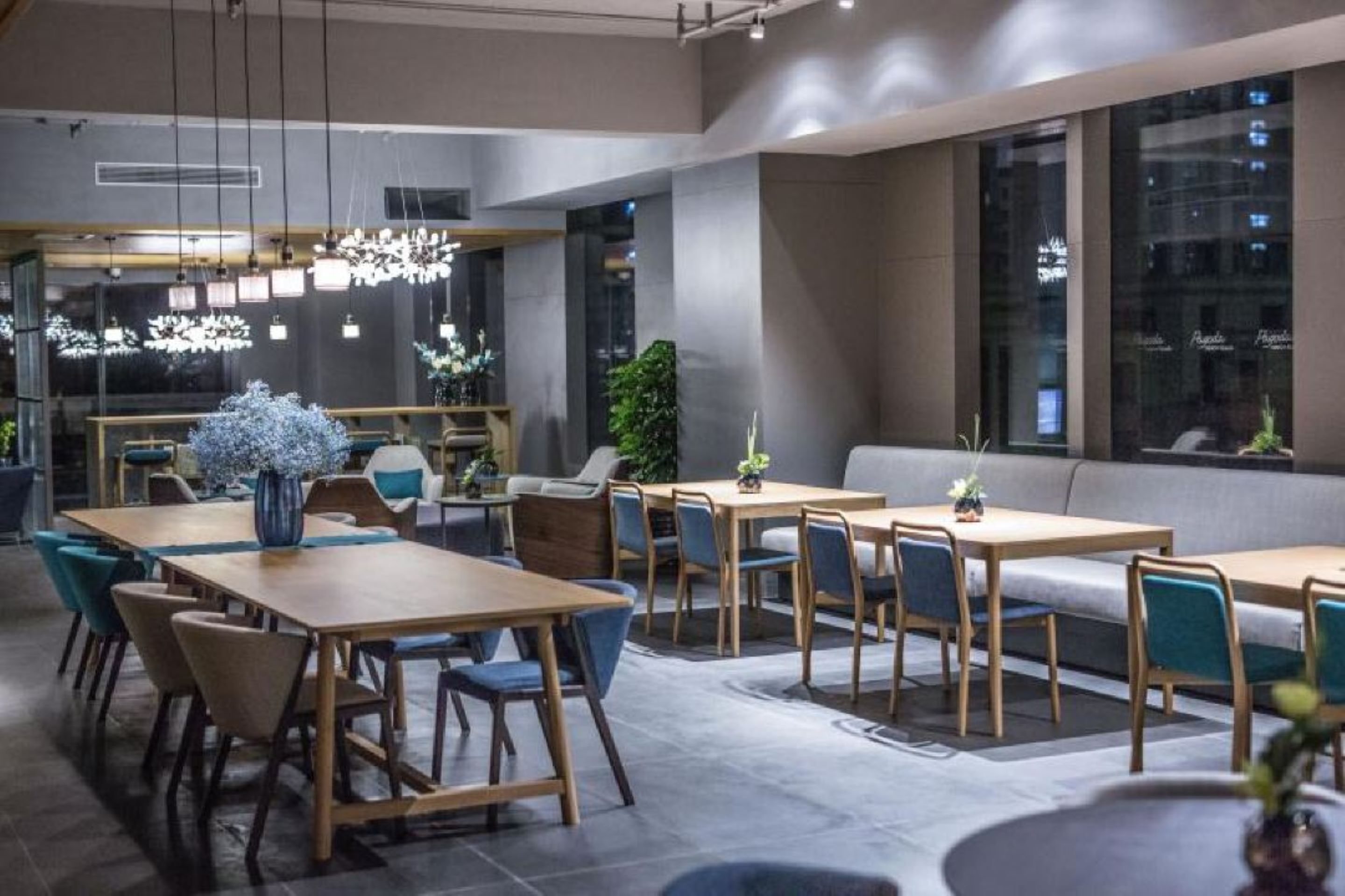 A modern dining area with wooden tables and blue chairs, large windows, and decorative plants.