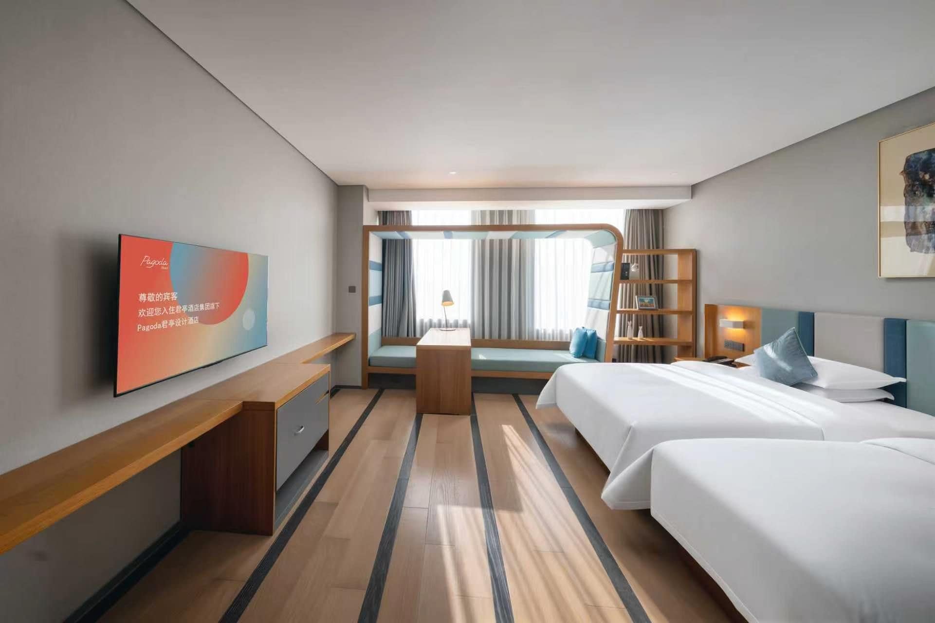 Una habitación de hotel con cama, mesitas de noche, escritorio, televisor, estantería y ventana con cortinas.