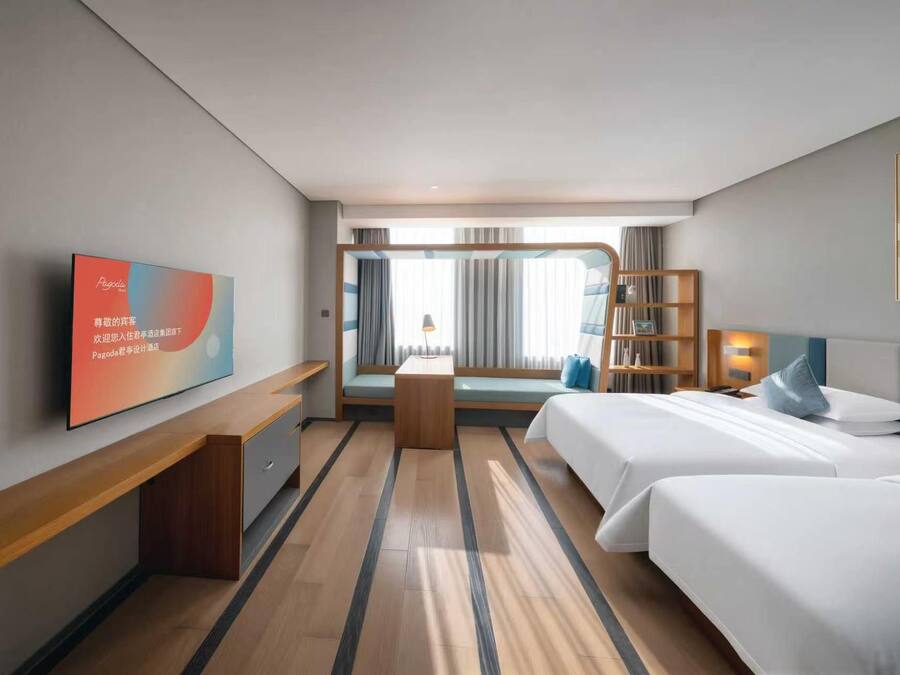 Una habitación de hotel con cama, mesitas de noche, escritorio, televisor, estantería y ventana con cortinas.