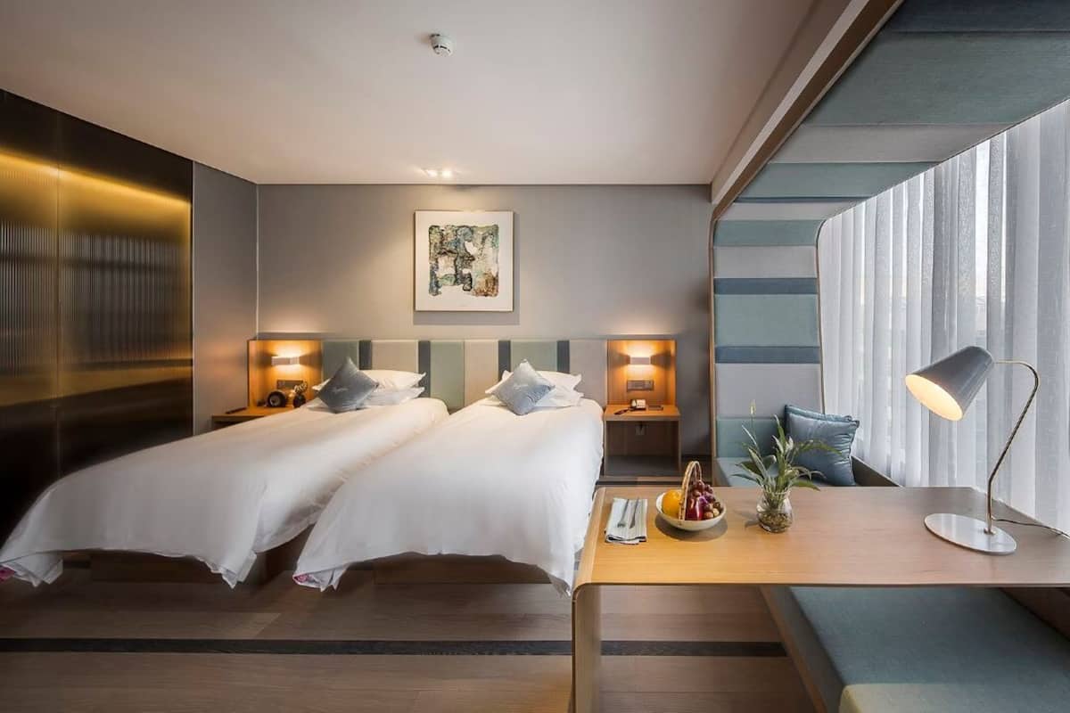Ein Hotelzimmer mit zwei Betten, einem Nachttisch mit Lampe, einem kleinen Tisch mit Obst und einem großen Fenster mit Vorhängen.