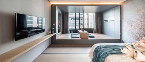A modern hotel room with a flat-screen TV mounted on the wall, a bed with a green and white striped bedspread, a seating area with a bench and a small table, and a view of the city through large windows.
