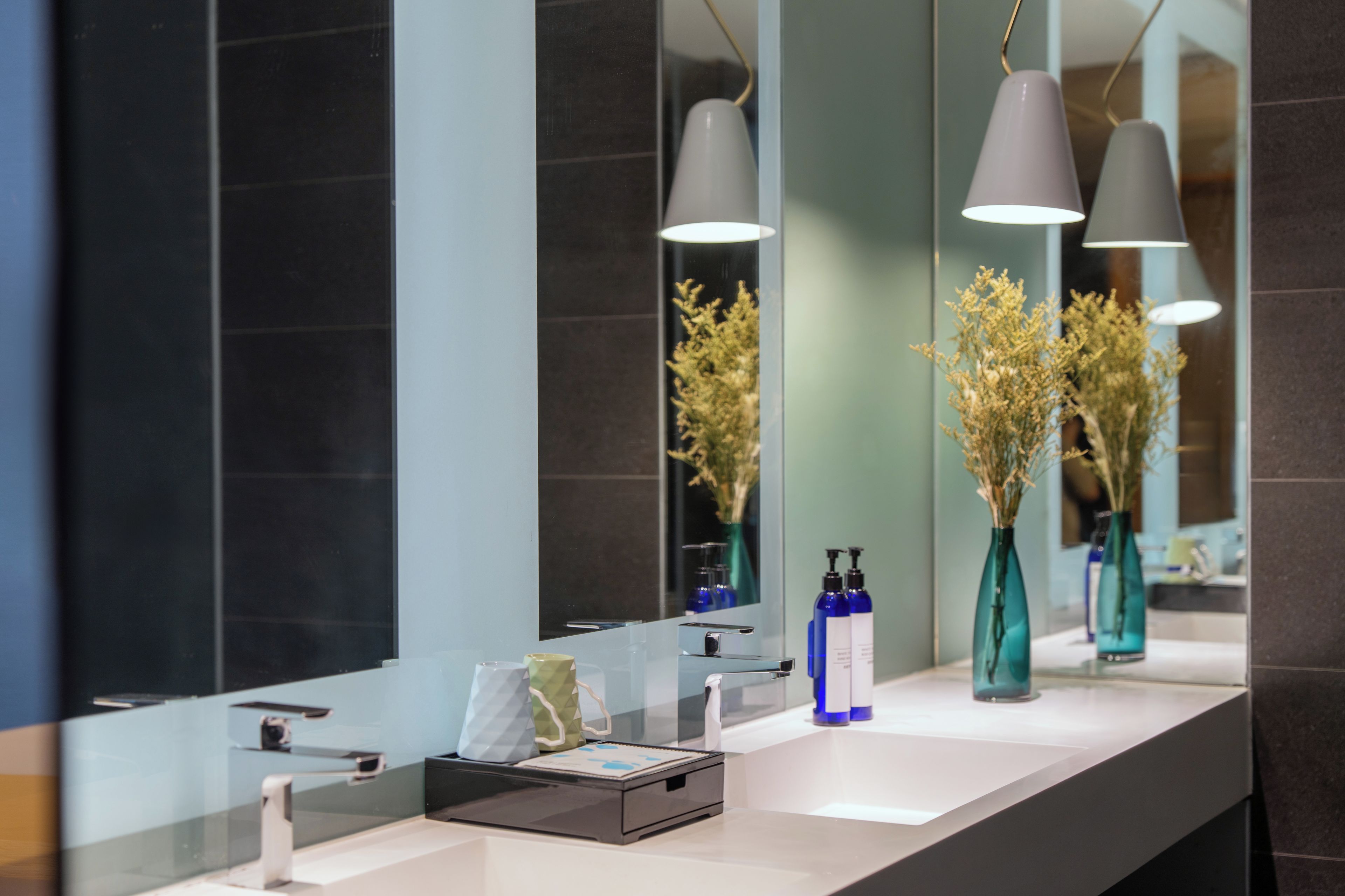 A modern bathroom with a large mirror, two pendant lights, and a vase with dried flowers.