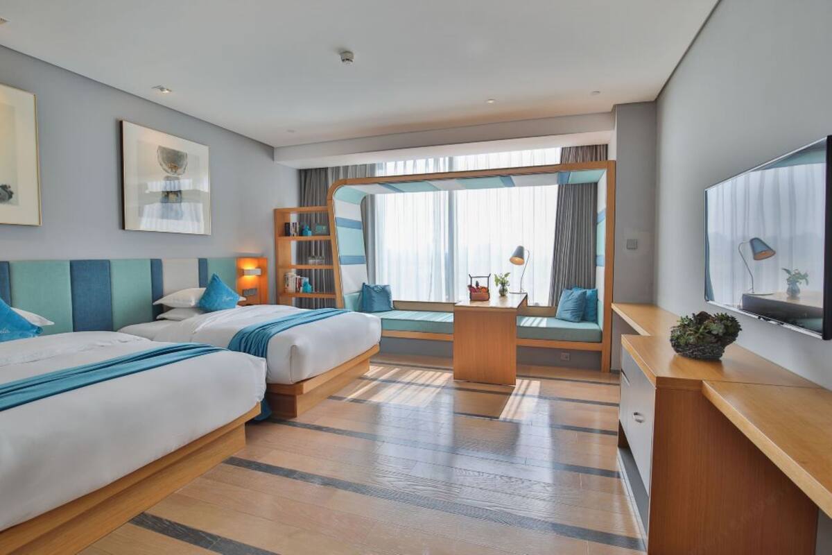 Ein Hotelzimmer mit zwei Betten, einem Schreibtisch, einem Fernseher und einem großen Fenster mit Vorhängen.