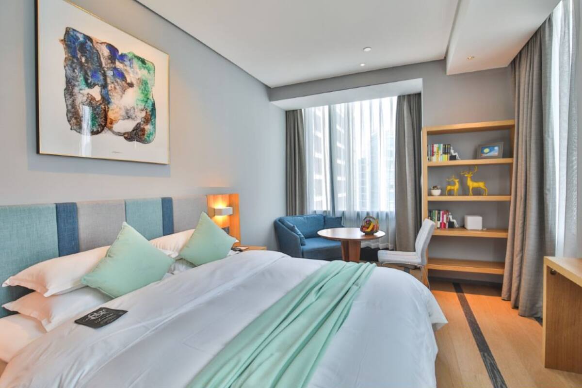 Ein Hotelzimmer mit Bett, blauem Sofa, kleinem Tisch, Bücherregal und großem Fenster mit Vorhängen.