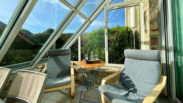 Una veranda con tetto in vetro, due poltrone grigie rivestite in tessuto, un piccolo tavolino in legno e un muro in pietra.