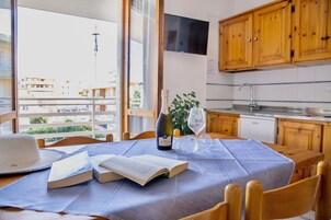 Un comedor con una mesa dispuesta para dos personas, que incluye un libro, una botella de vino y una copa. La habitación tiene armarios de madera, un ventanal con vista a edificios y una zona de cocina con un fregadero y alacenas.