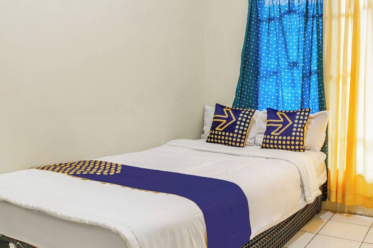 Una cama bien tendida con ropa de cama blanca y almohadas estampadas en azul y amarillo, una manta de color azul y amarillo y una cortina azul con lunares blancos.