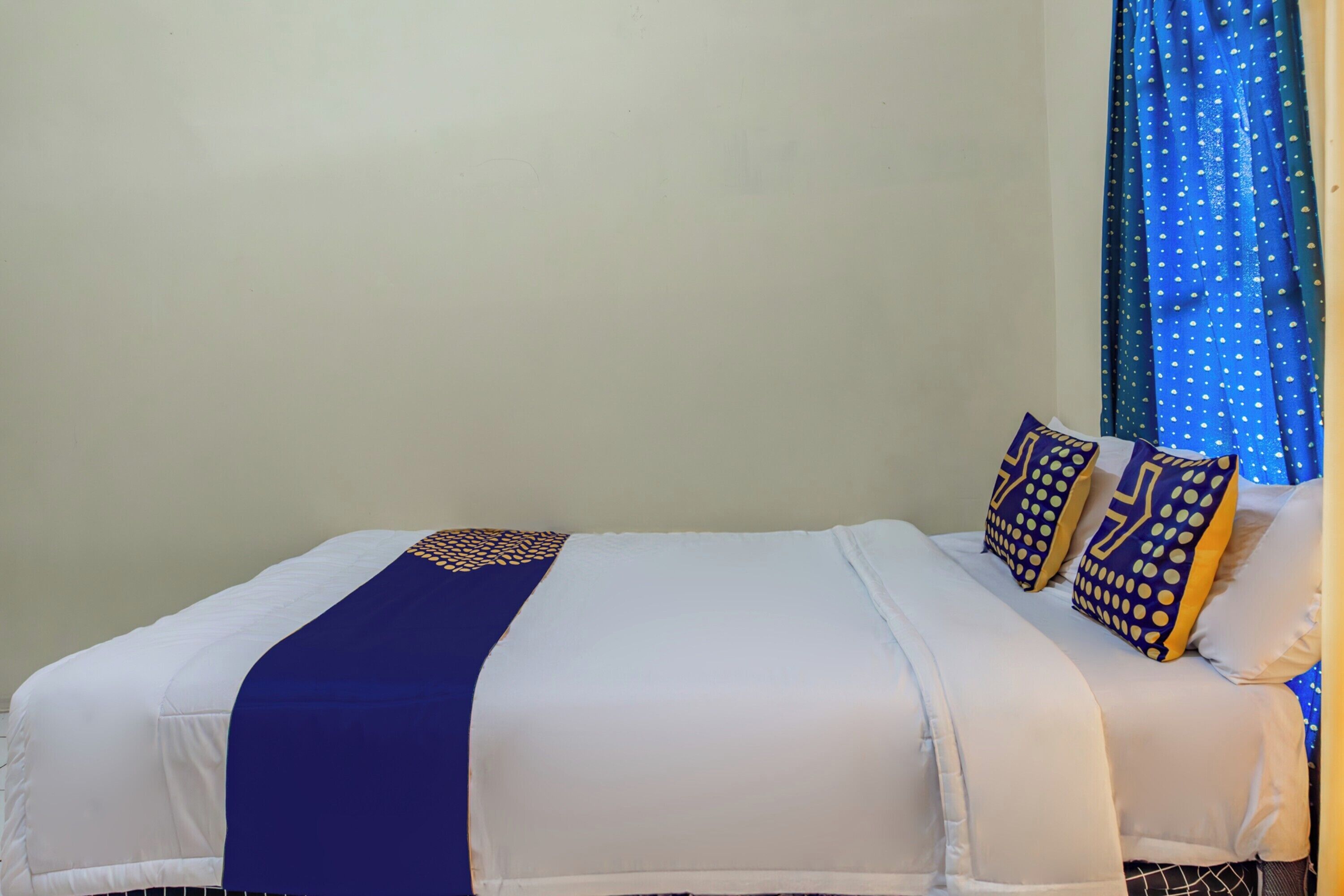 Un lit bien fait, recouvert d’une couvre-lit bleue et blanche assortie aux coussins, un mur sobre et une fenêtre ornée de rideaux bleus.
