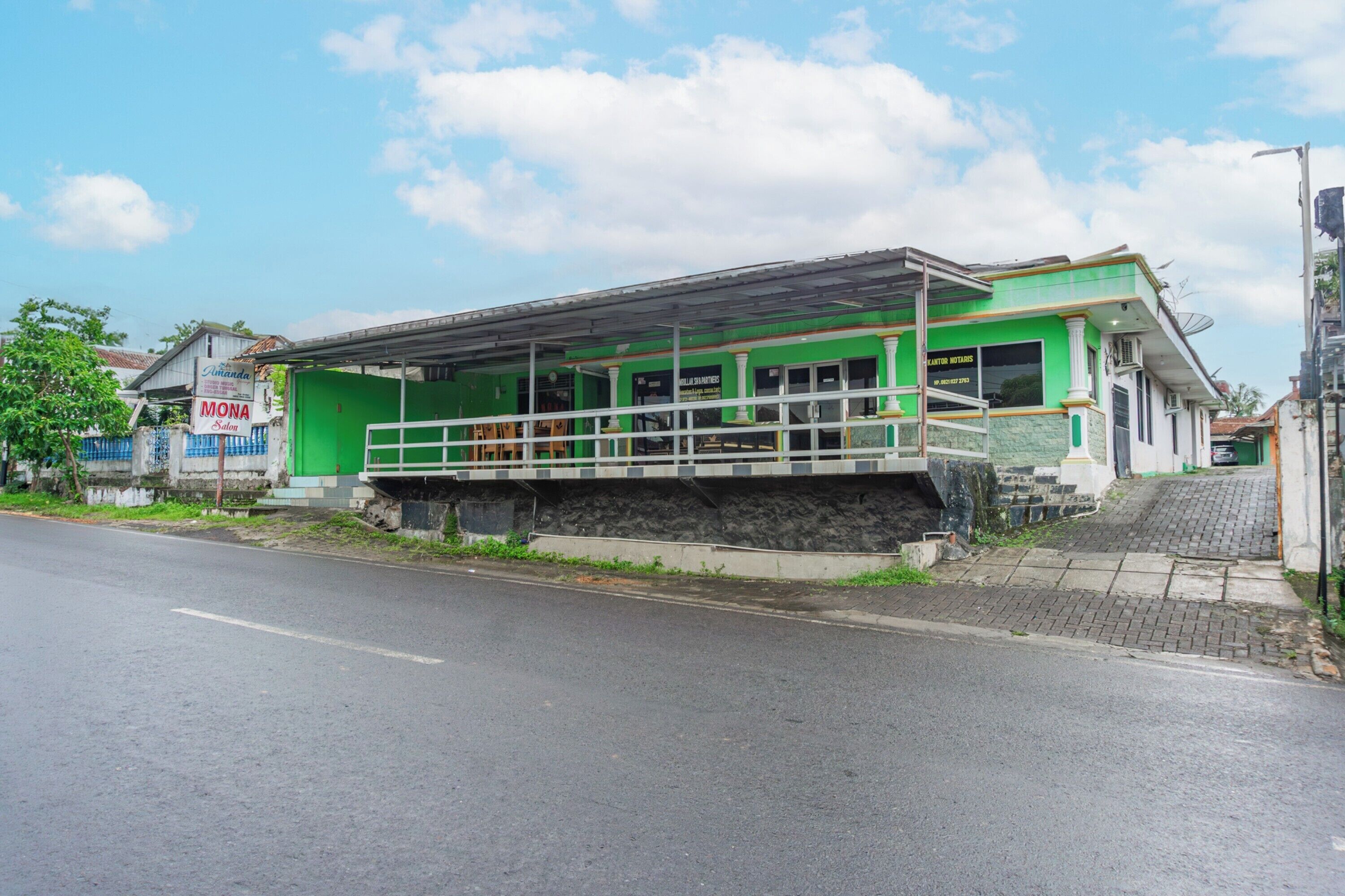 A green building with a covered outdoor area, a visible entrance, and a paved sidewalk.