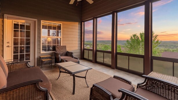 Un espacio al aire libre techado con mobiliario de mimbre, ventilador de techo y amplios ventanales con vistas al paisaje.