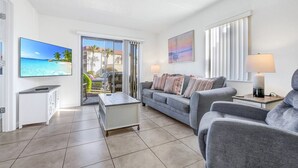 Ein modernes Wohnzimmer mit grauem Sofa, Couchtisch und Fernseher. Eine Glastür öffnet sich zu einem Balkon mit Blick auf Palmen und ein Auto.
