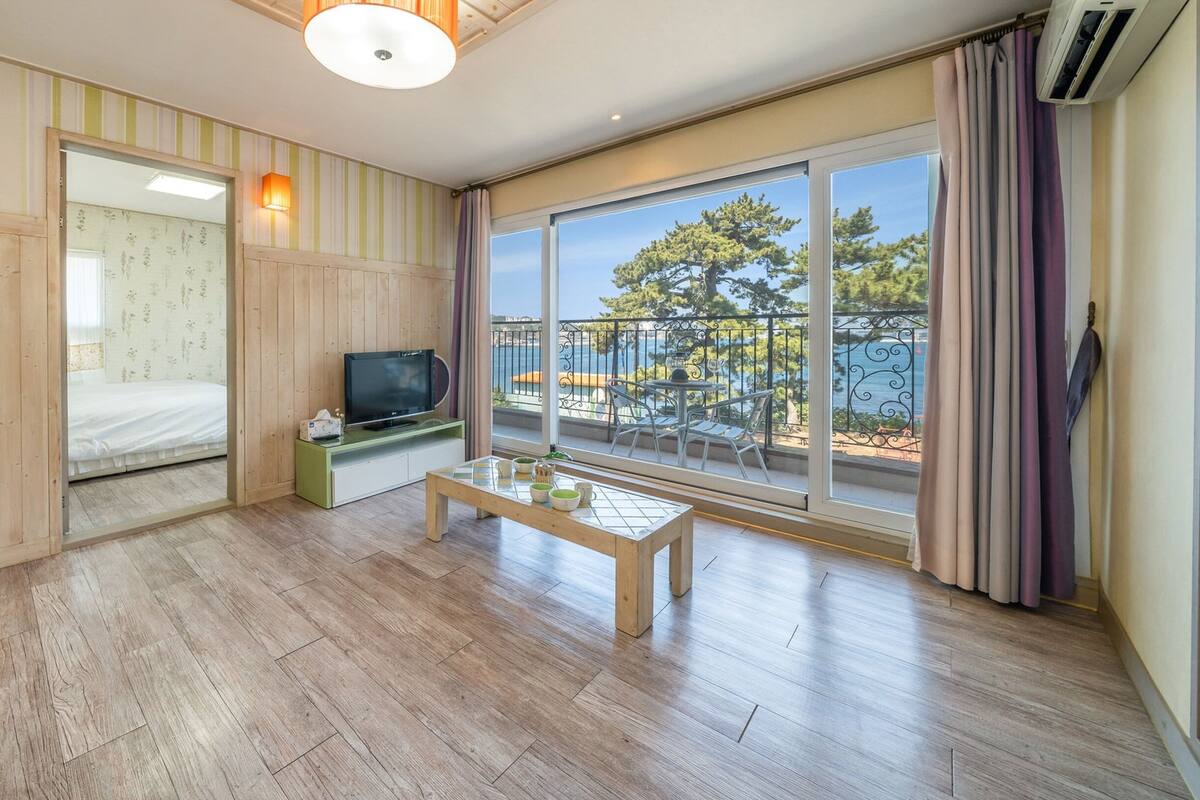 Una habitación con piso de madera, un televisor sobre un soporte, una mesa con tazas y un control remoto, una ventana con vista a árboles y un balcón, y una pared a rayas.