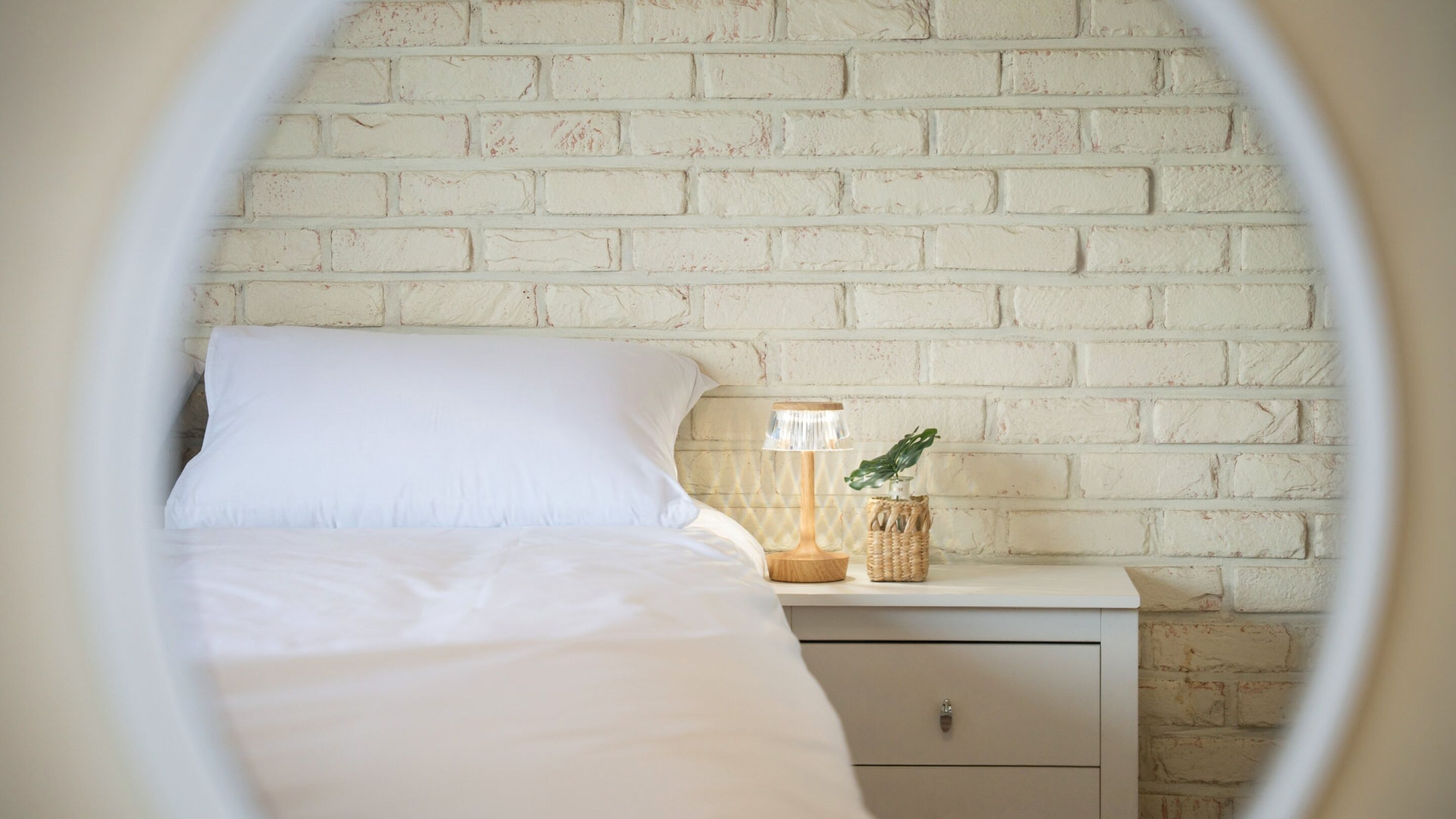 Ein Bett mit weißer Bettwäsche, ein Nachttisch mit Lampe, eine kleine Pflanze und eine weiße Backsteinwand.