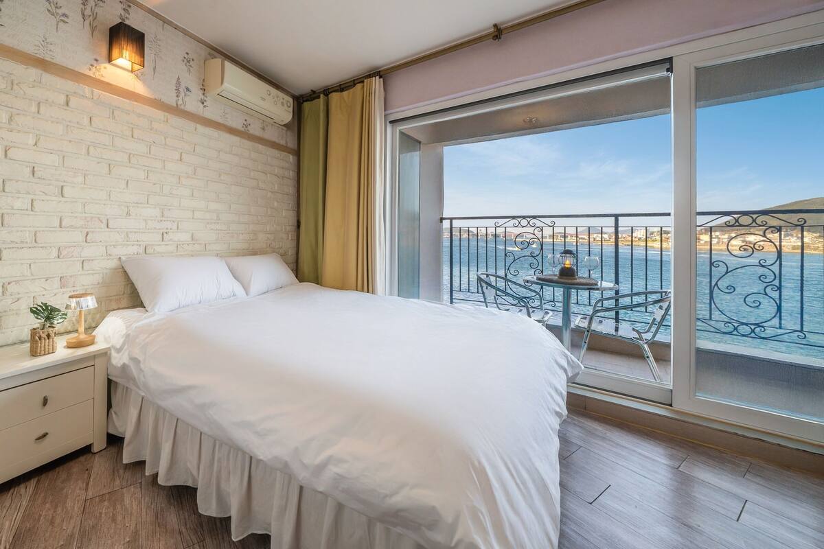 Un dormitorio con una cama grande, una mesita de noche, una lámpara y vistas al mar desde un balcón.