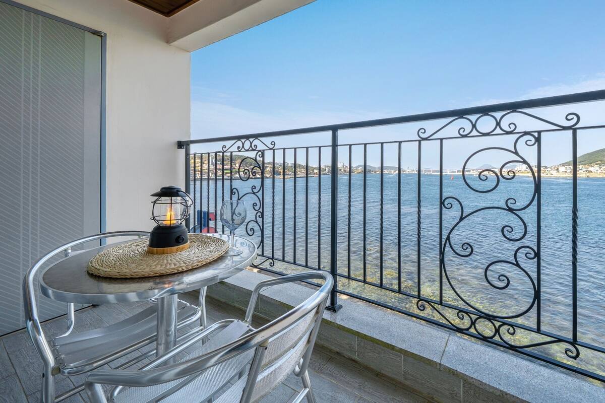 Un balcón con mesa y sillas, un farol y vista al mar.