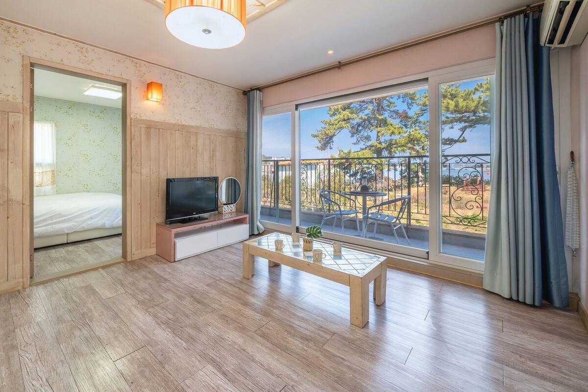 Una habitación con piso de madera, un televisor sobre un mueble bajo, una mesa de vidrio con tazas, un balcón con vistas a árboles y una cerca, y una puerta corrediza que da a otra habitación.