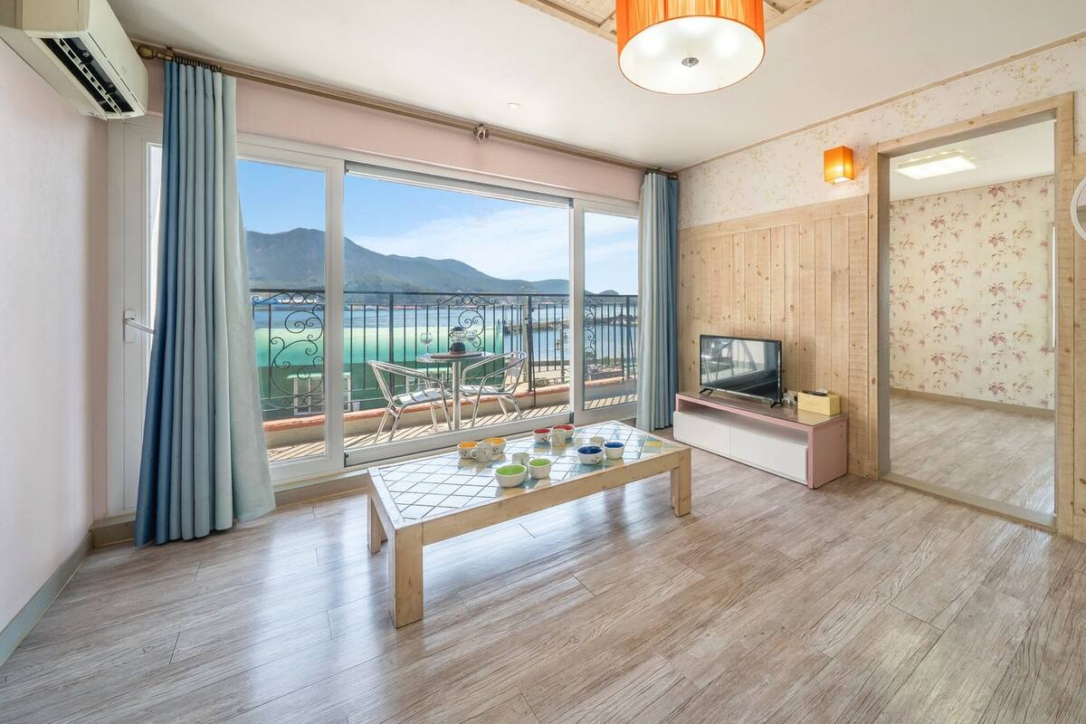 Habitación con un gran ventanal corredizo que da a un balcón con vistas a montañas y un cuerpo de agua. El suelo es de madera, hay una mesita con cuencos, y un televisor sobre un mueble.