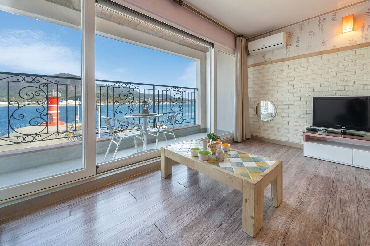 Un balcón con vistas al mar, una mesa con cuencos y un televisor sobre un mueble.