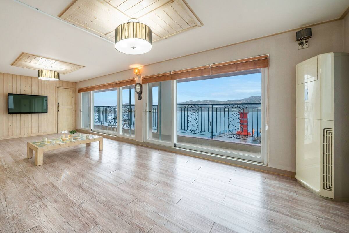 Amplia sala de estar con piso de madera, un ventanal con vista al mar, un televisor de pantalla plana montado en la pared y una lámpara de techo moderna.