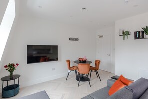 Ein modernes Wohnzimmer mit einem Flachbildfernseher, einem Esstisch mit Stühlen, einem grauen Sofa und einem kleinen Beistelltisch mit Blumen.