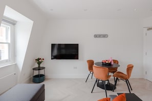 A modern room with a flat-screen TV mounted on the wall, a dining table with chairs, a small side table with a vase, and a sign that reads 'NO RIF RAFF'.
