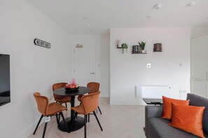 A modern living room with a dining area, a sofa with orange cushions, a flat-screen TV, and a 'NO RUFF' sign on the wall.