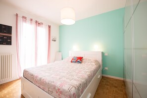 A bedroom with a bed, a bedside table, a wall-mounted light, a radiator, and a window with curtains.