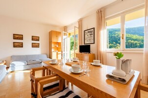Un comedor con mesa de madera, sillas y televisor. Hay cuadros en la pared y una ventana con vistas a árboles.