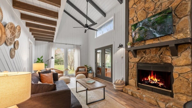 A spacious living room with a stone fireplace, a ceiling fan, and a large window overlooking a garden.
