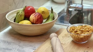 Una encimera de cocina con un cuenco de fruta, un tazón de cereales y una tabla de cortar de madera.