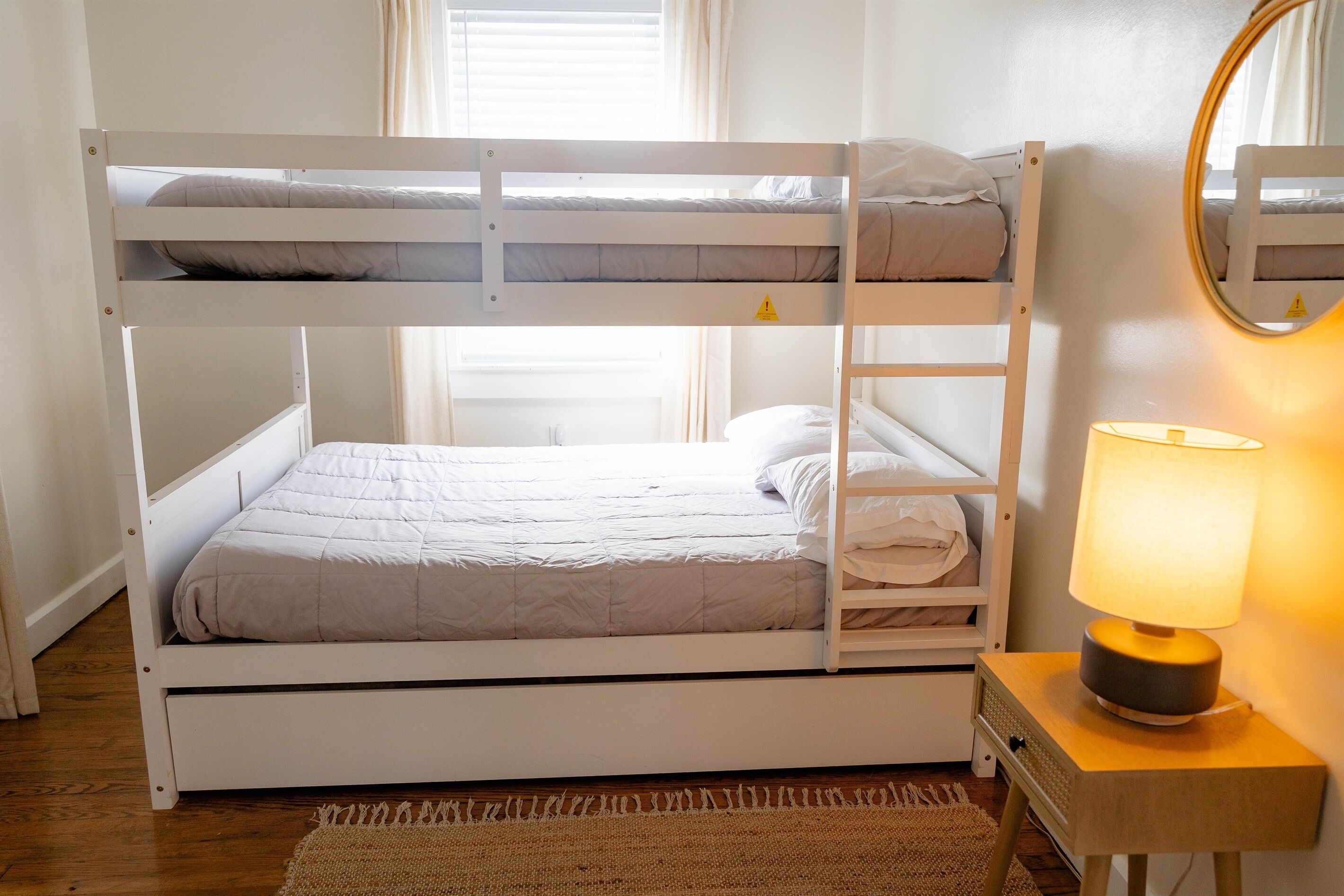Een stapelbed met nachtkastje en lamp, een spiegel en een raam met gordijnen.