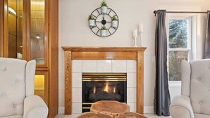 Un salon avec une cheminée, deux fauteuils, une horloge murale ronde et une fenêtre avec des rideaux.