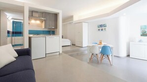 Una sala de estar moderna y bien iluminada, con un sofá, una mesa de comedor con sillas, una kitchenette y una cama.