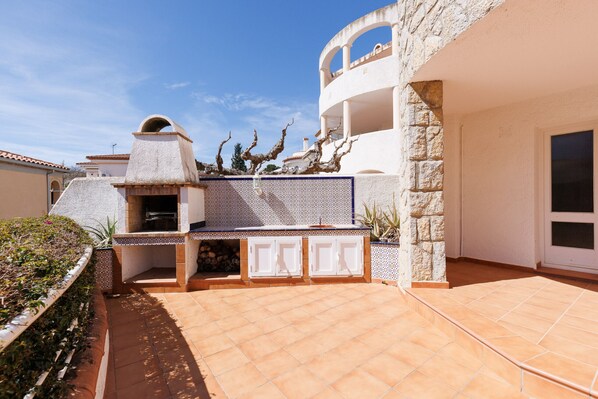 Un espace patio aménagé avec un barbecue, un mur en pierre et un bâtiment blanc doté d’un balcon.