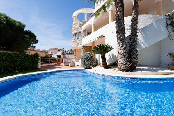 A swimming pool with a clear blue water surface, surrounded by a white building with a stone facade and multiple balconies.