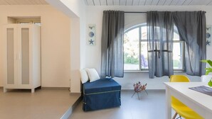 Una habitación con cama, escritorio y silla. Se observa una ventana con cortinas, una planta y una pequeña mesa con un objeto decorativo.