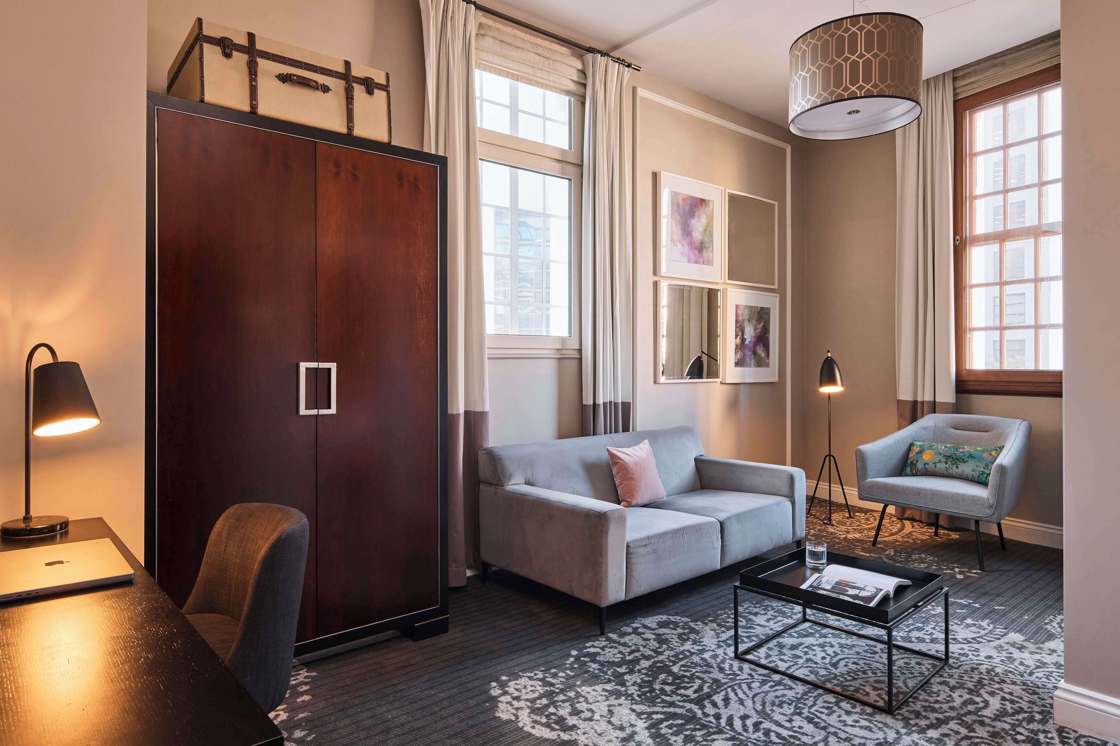 A modern hotel room with a grey sofa, a small table, a desk with a lamp, a chair, and a large wardrobe.