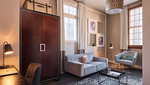 A modern hotel room with a grey sofa, a small table, a desk with a lamp, a chair, and a large wardrobe. - The Inchcolm by Ode Hotels (Spring Hill)