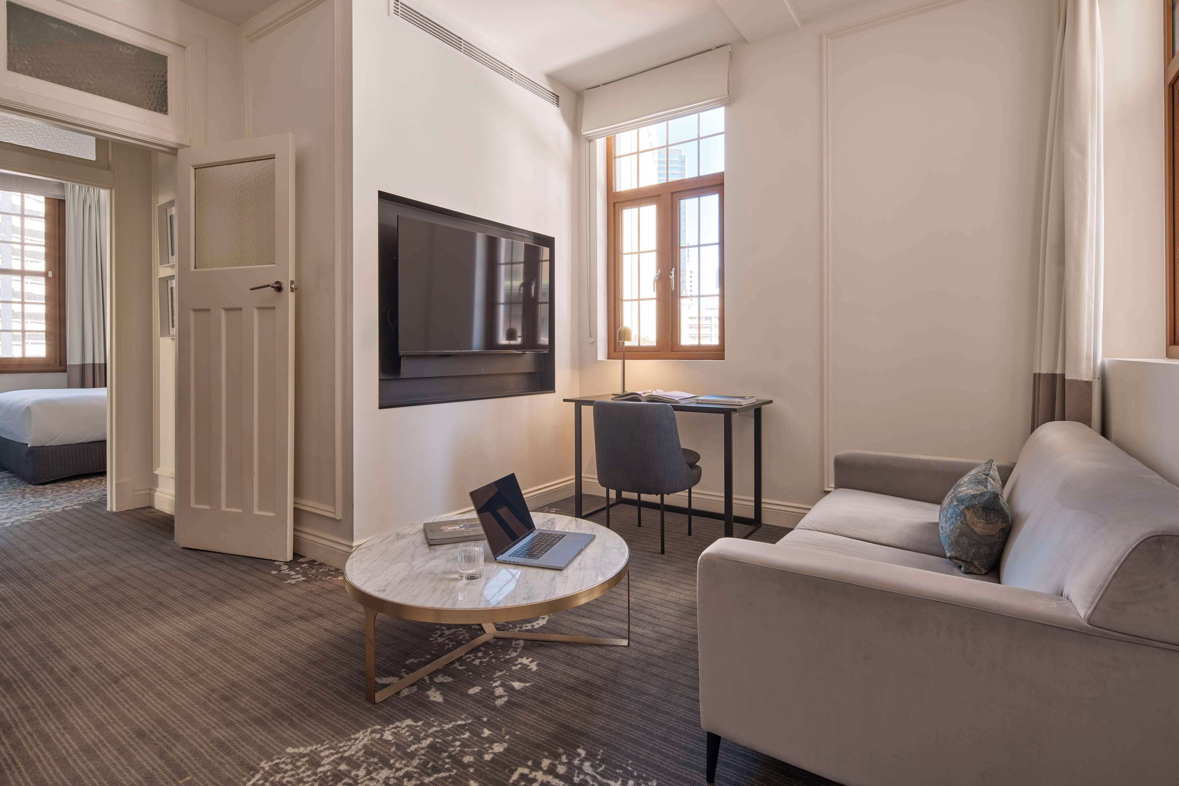 A hotel room with a bed, a round table with a laptop, a sofa, a TV, and a window with curtains.
