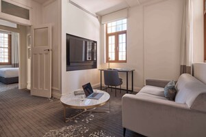 Habitación de hotel con una cama, una mesa redonda con una computadora portátil, un sofá, un televisor y un ventanal con cortinas.