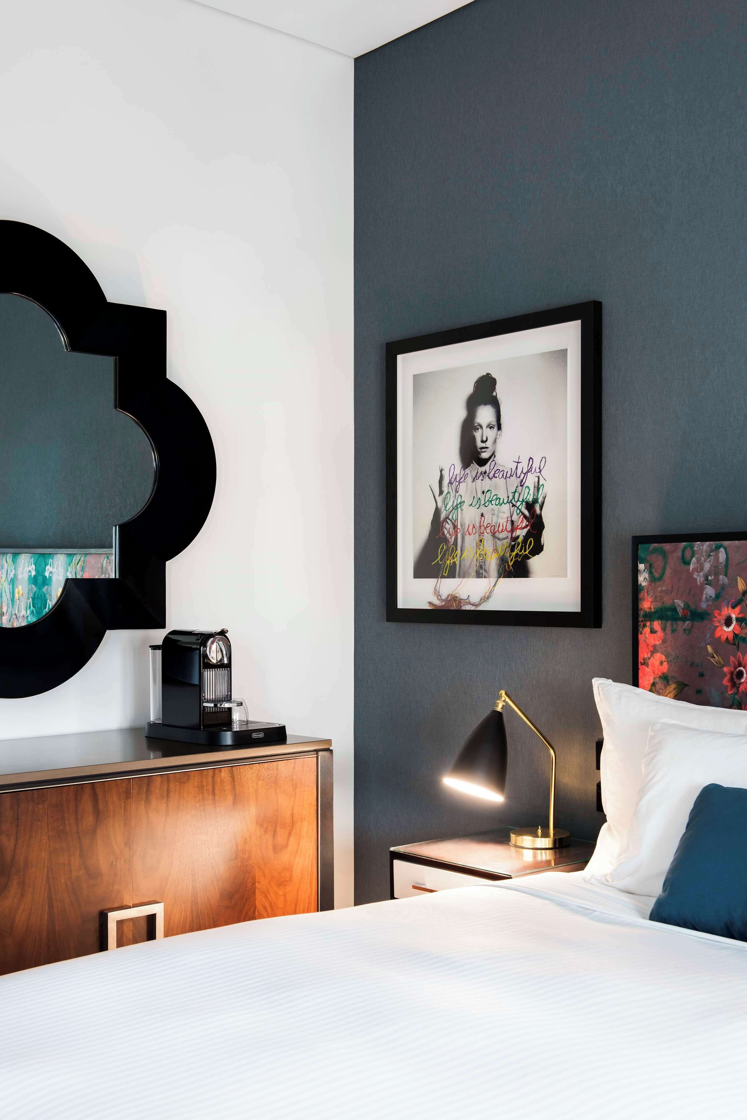 A modern hotel room with a bed, a bedside table with a lamp, a coffee maker, and a framed quote on the wall.